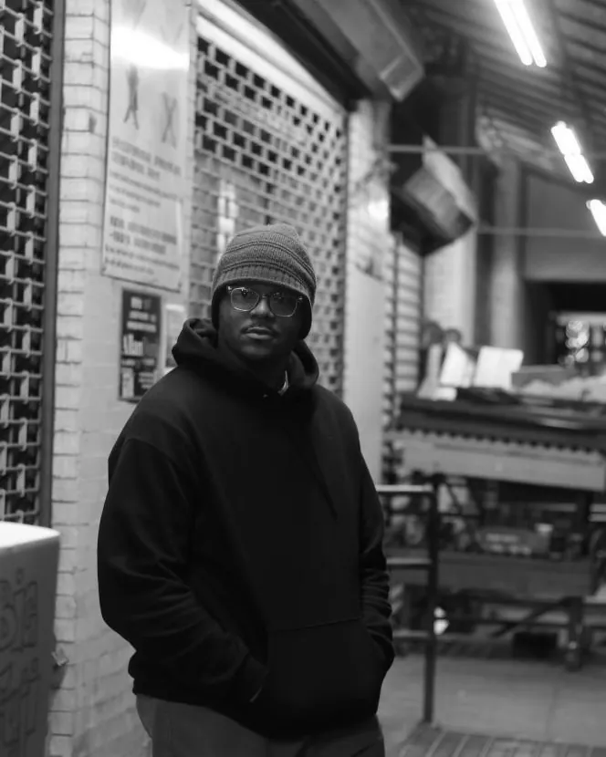 Stonzyx in a beanie and hoodie under the lights of a shuttered storefront